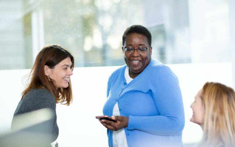 A group of three women in an office setting, chatting and laughing with each oth